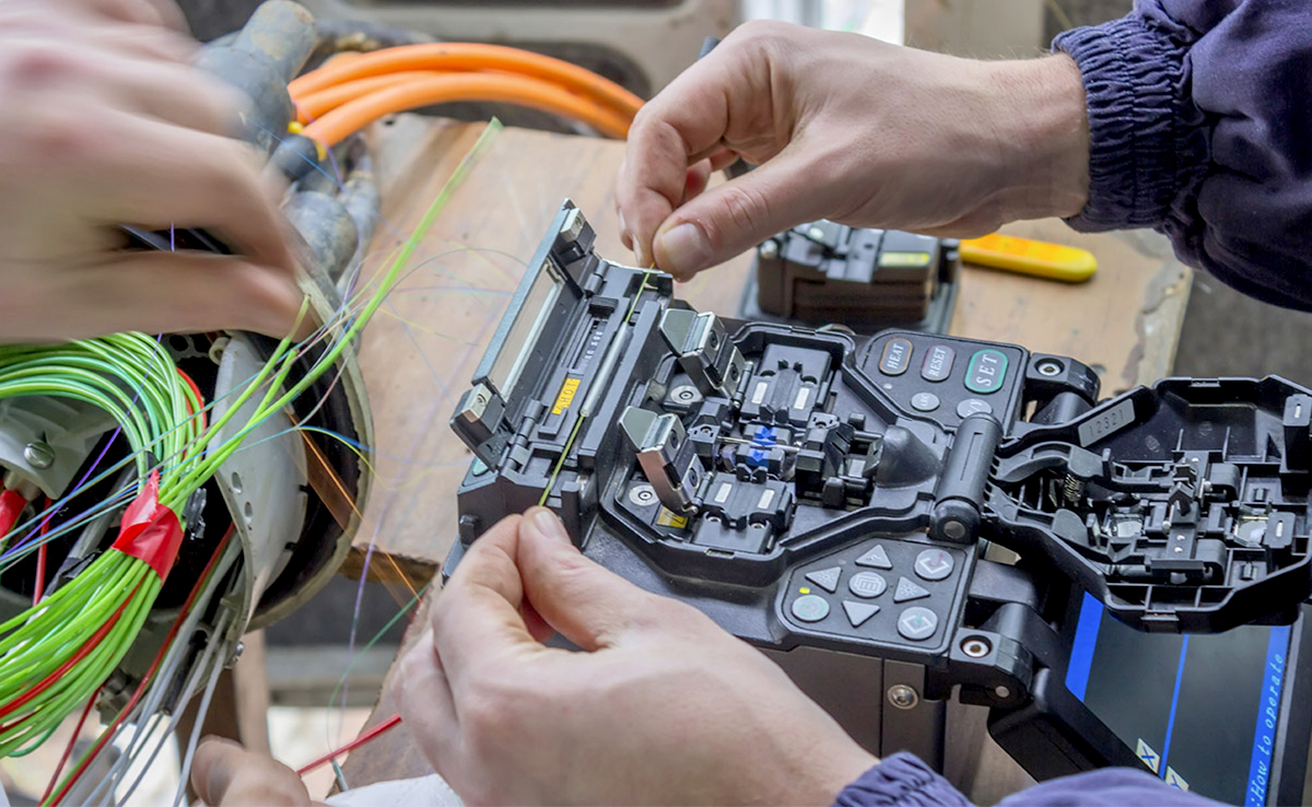 Dois técnicos posicionando fibras ópticas em máquina de fusão por arco elétrico sobre bancada