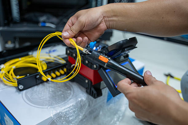 Mãos segurando cordão óptico amarelo com conector diante de ferramenta de conectorização sobre bancada
